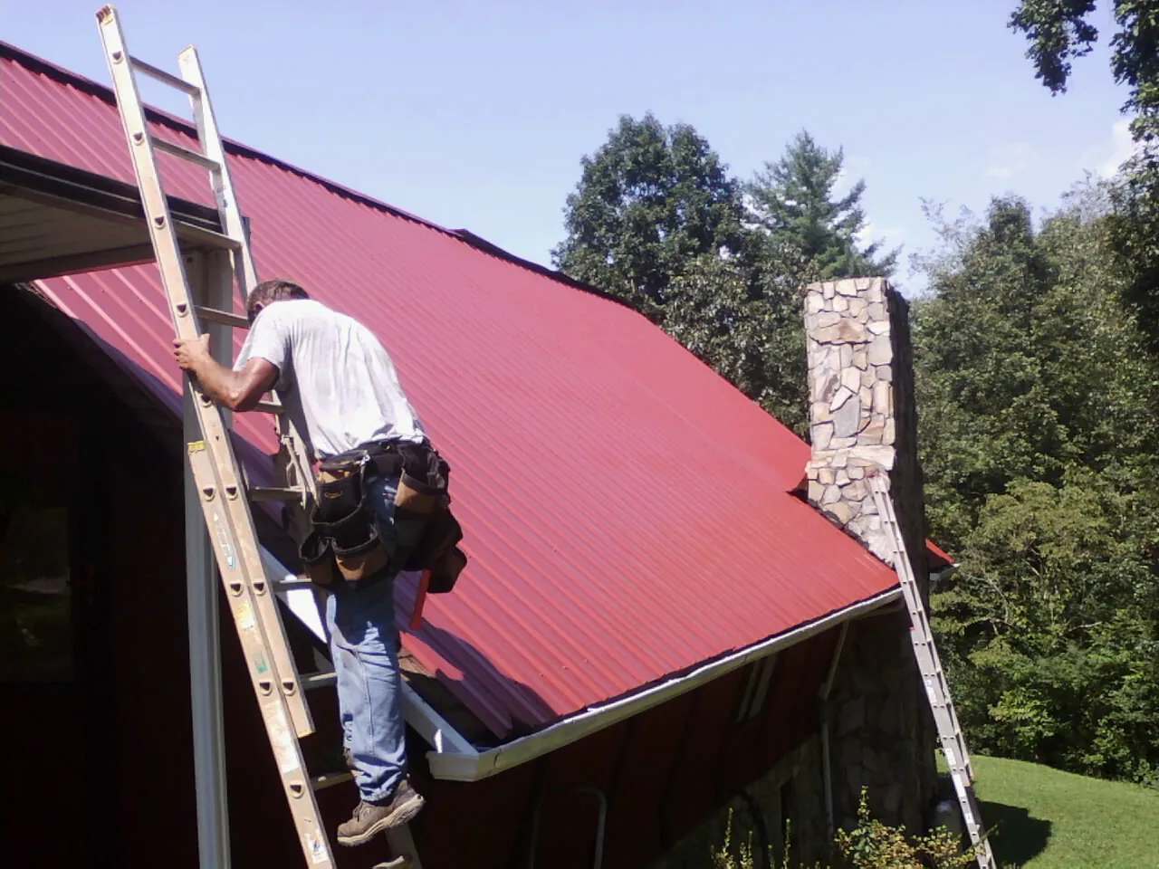 Roofer on a ladder beside a red metal roof and stone chimney — Soffit & Fascia Repair by Roofing Brothers in Scales Mound